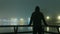 A hooded figure silhouetted against street lights next to a river. On a mysterious misty, winters night