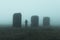 A hooded figure standing by mysterious abandoned industrial pillars, standing on moorland. On a spooky, foggy day