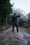 A hooded man, back to camera on a muddy path holding a spade, next to a log pile on a grey moody day in the countryside