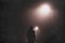 A hooded man standing underneath a street light, back to camera. On a foggy winters night. With a blurred, textured edit