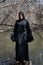 Hooded woman in black robes standing on a tree stump in the middle of a swamp. Halloween and Gothic concept. Witchcraft and magic