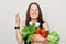 Hopeful adult woman with brown hair standing isolated over white background holding vegetables crossing her finger closing eyes