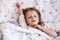 Horizontal indoor portrait of lovely charming little kid raising one hand, having peaceful facial expression, lying in bed,