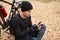 Horizontal outdoor picture of young persistent sportsman sitting near his bicycle in forest, typing messages, using his phone,