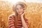 Horizontal outdoor shot of pretty charismatic model sitting in wheat field, having peaceful facial expression, touching her chin