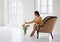 Horizontal photo of a girl in a beige dress who is sitting in a chic armchair in a white studio