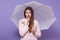 Horizontal photo of thoughtful attentive attractive female holding umbrella, putting one finger on cheeck, having pensive facial