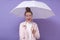 Horizontal portrait of magnetic adorable young female with bun holding light umbrella, covering herself from rain, opening mouth