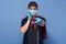 Horizontal shot of astonished male wearing t shirt, sweater over shoulders, cap, protective mask,latex gloves, courier hands black