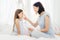 Horizontal shot of attractive brunette female mother applies cream on nose of her daughter, sit together on bed in bedroom, have g