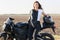 Horizontal shot of attractive motorrcyclist leans at motorrbike, keeps hand on helmet, stands near bike, has leather jacket on her