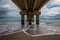 Horizontal shot of a beach under a pier with waves splashing against the pier`s columns
