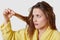 Horizontal shot of beautiful dark hair woman looks at her wet damaged hair, has stressful facial expression, wears yellow bathrobe