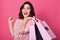 Horizontal shot of brunette woman, looks happy, stands laughing and holding shopping bags, posing isolated over pink studio