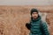 Horizontal shot of contemplative good looking man has stubble, wears hat, jacket and gloves, stands near wheat field background