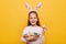 Horizontal shot of cute optimistic little girl wearing rabbit ears holding Easter eggs in wicker basket, showing testicle painted