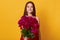 Horizontal shot of cute young woman with flowers standing isolated over yellow background, female giving bouquet of maroon peonies