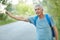 Horizontal shot of handsome grey haired male in casual clothes, hitchhikes on road, carries backpack, has journey in countryside,