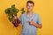 Horizontal shot of handsome model standing isolated over yellow background in gray t shirt, holding raw vegetable in hand, smiling