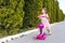 Horizontal shot of happy pretty little girl wearing pink dress and backapack on the scooter against the green trees on the street