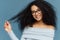 Horizontal shot of lovely smiling Afro American woman holds tips of her curly hair, wears fashionable light blue sweater,