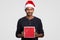 Horizontal shot of pleasant looking male holds red present, wears festive hat, recieves present on New Year or Christmas, models