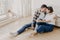 Horizontal shot of pleased husband and wife embrace and kiss, have good relationship, sit on wooden floor near couch, enjoy being