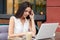 Horizontal shot of pretty busy female freelancer sits in front of opened laptop, drinks coffee, works on new project, surrounded w