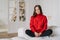 Horizontal shot of relaxed housewife dressed in warm red knitted sweater, sits crossed legs in lotus pose on couch, in spaciouss r