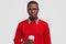 Horizontal shot of serious dark skinned man holds disposable cup of coffee, dressed in red jumper, looks confidently at