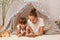 Horizontal shot of woman and her daughter laying in peetee tent and watching cartoons on mobile phone, spending time together at