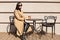 Horizontal shot of young woman with cup drinks cocoa at city street cafe terrace, sits at table and dreaming, lady wears beige