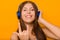 Horn gesture close up. A young girl listens to music with headphones, standing on a yellow background.