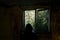 A horror concept. Of a hooded figure, back to camera. Looking out of a broken window. In an abandoned spooky ruined house