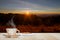 Hot coffee cup and biscuit on wooden table top on blurred meadow and mountain with sunrise and flare background