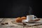 Hot coffee with steam in a white cup with muffins, cinnamon and star anise on burlap on a wooden table on a black concrete