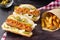 Hot dogs with vegetables, mustard and ketchup on a cutting board on a dark background. French fries and gherkins, side view