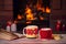 Hot drinks in mugs, book and candles on wooden table beside cosy open fire place. Autumn or winter holidays concept.