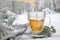 Hot herbal tea is poured in a glass cup outdoors in the snow, beside some sage leaves and a knitted scarf, medicinal herbs and