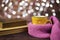 Hot tea, hot chocolate, coffee in yellow cup, wrapped with a pink knitted scarf. Old books. Blurred lights, wooden background.