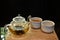 hot tea with tea leaves in glass teapot with ceramic cup on wood plate on glass table with selective focus on