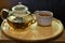 hot tea with tea leaves in glass teapot with ceramic cup on wood plate on glass table with selective focus on