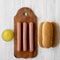 Hotdog ingredients: sausages on wooden board, hot-dog buns and mustard on white wooden surface, top view. Flat lay, overhead, from