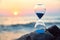 Hourglass with sand standing on rock. Sunset over sea and nature landscape. Running of time and relax idea, copy space