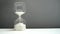 Hourglass on a white table and gray background. Faceted modern hourglass with light gray sand.