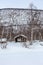 The house among the birches against the snow-covered field and the slope of a hill overgrown with trees.