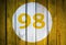 House number or calendar date in white circle on yellow toned wooden door background. Number ninety eight, 98