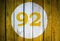 House number or calendar date in white circle on yellow toned wooden door background. Number ninety two 92