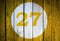 House number or calendar date in white circle on yellow toned wooden door background. Number twenty seven 27