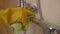 A housewife in protective rubber yellow gloves cleans a stainless steel faucet with a washing gel. Dirty bathroom faucet